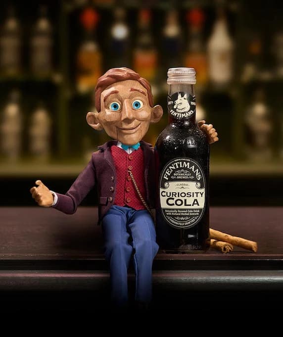 A puppet-like character sits beside a large bottle of Fentimans Curiosity Cola in a bar setting.